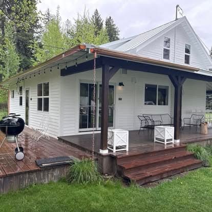 Charming white cottage with wooden deck, grill, and outdoor seating in a lush green setting.