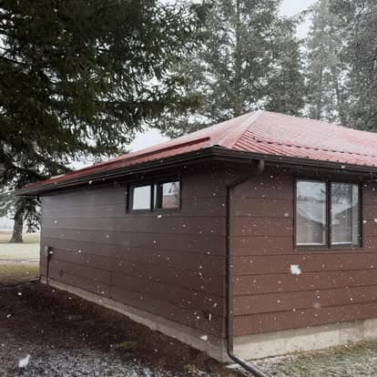 Snowy landscape featuring a modern brown house with a red roof amidst tall evergreen trees.