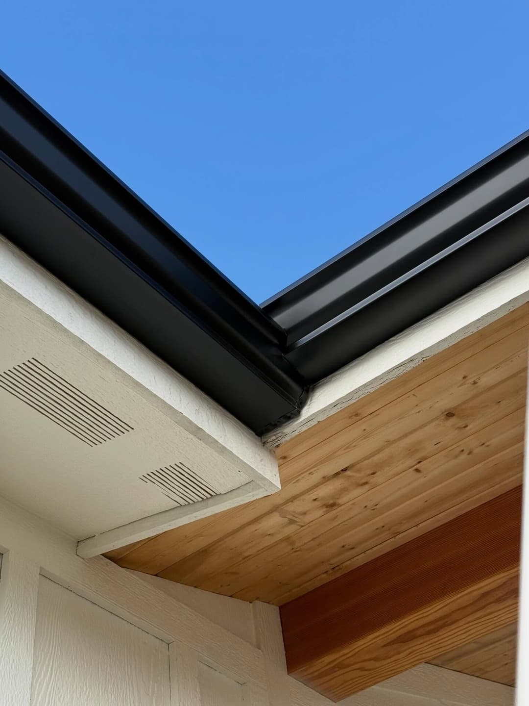 Roof corner showing black gutter, wooden overhang, and clear blue sky.