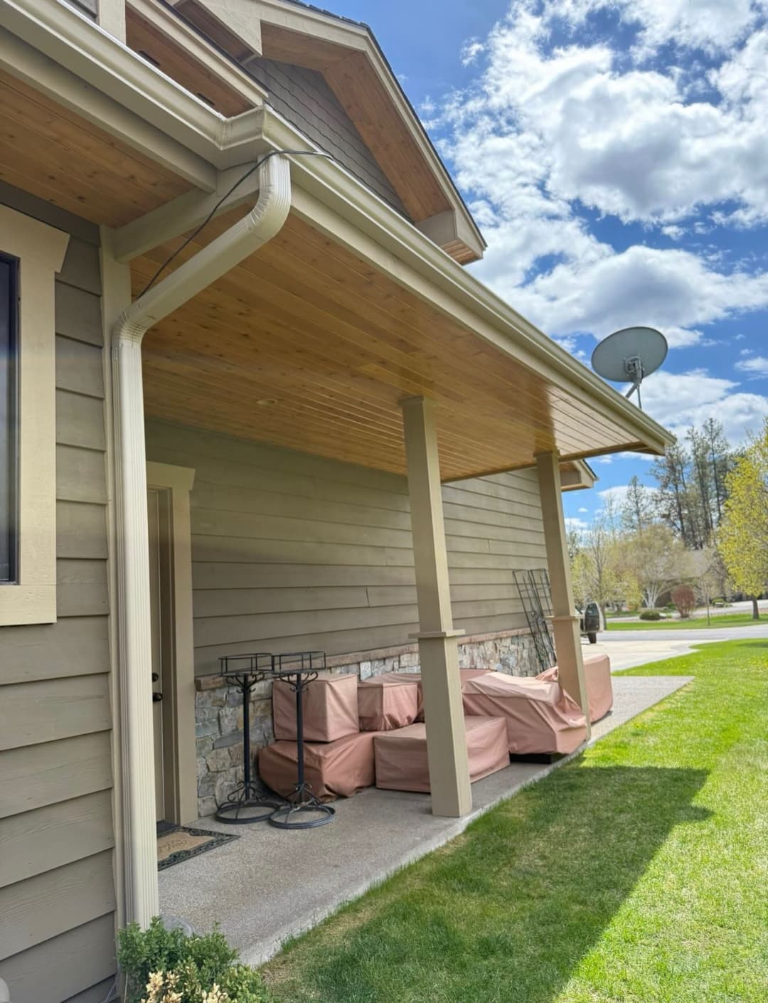 Front porch of a house with outdoor furniture covered, surrounded by green grass and blue sky.