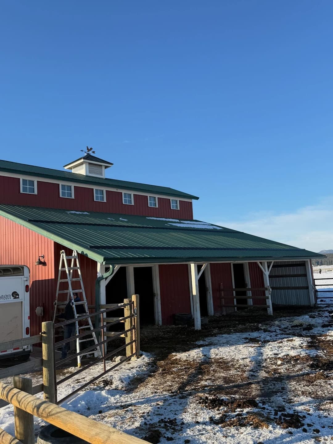 Red barn with green roof and solar panels, surrounded by snow and blue sky.