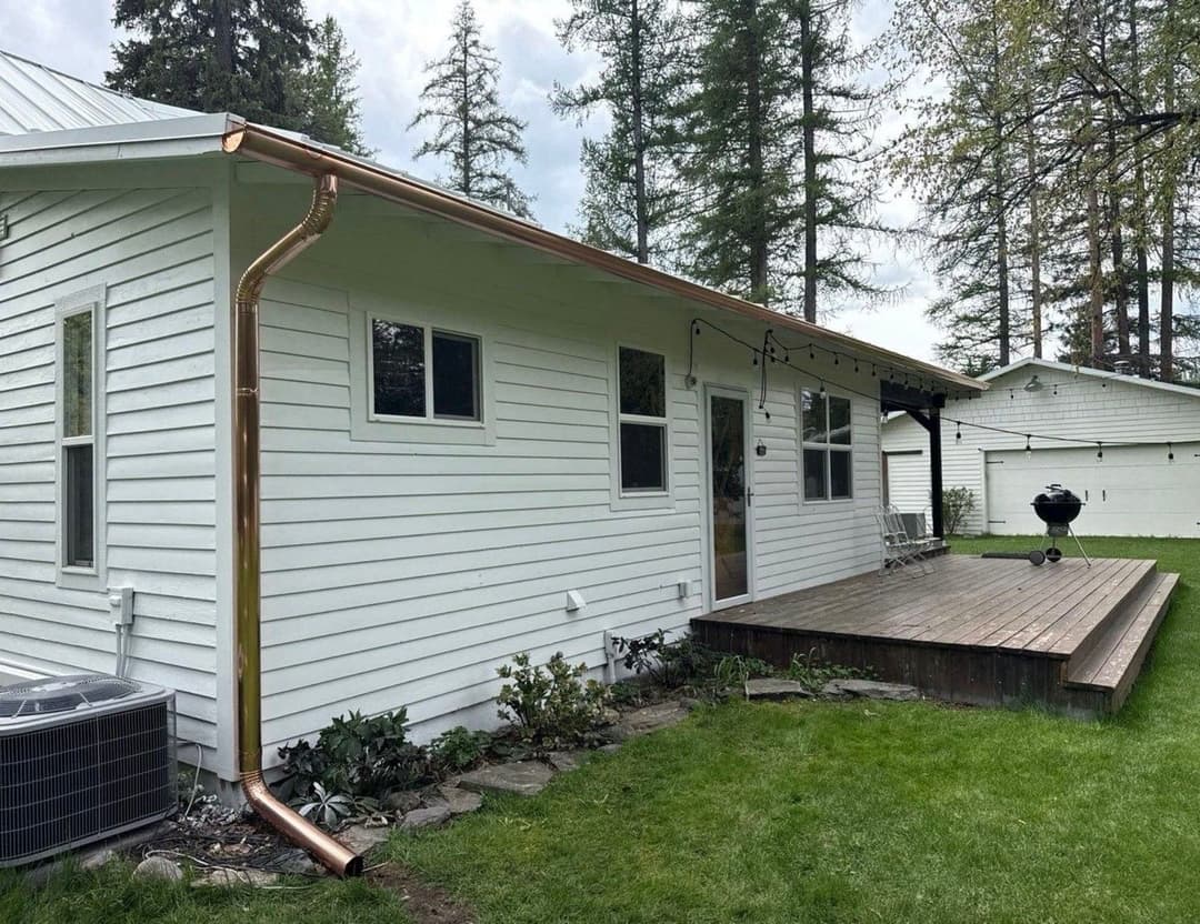 White house exterior with copper downspout, wooden deck, and green lawn in a tranquil setting.